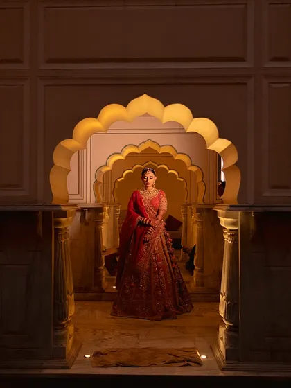 A bride stands framed by the magnificent scalloped arches of Alila Fort Bishangarh. The architectural beauty of the heritage venue, combined with soft lighting, creates a truly royal portrait.