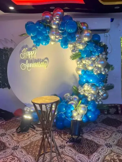 A simple yet elegant anniversary setup at home. A white circular backdrop with 'Happy Anniversary' text is accented by a blue and silver balloon garland with integrated fairy lights.