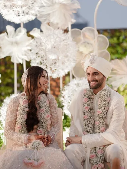 A moment of shared laughter during the ceremony. The couple, dressed in elegant white, look at each other with genuine happiness, seated against a stunning floral backdrop.