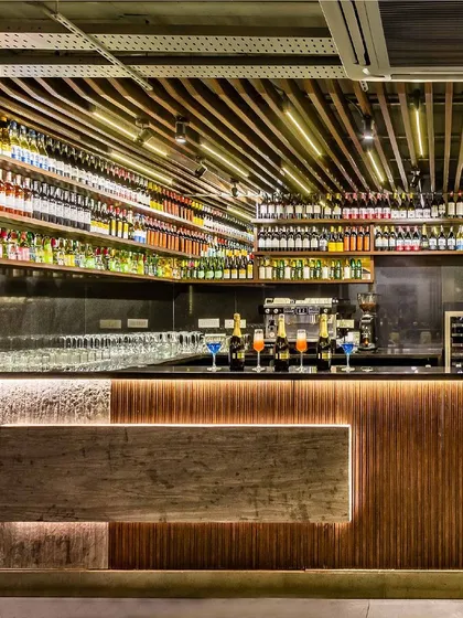 The exquisite bar counter at Pasta Street, crafted with a mix of stone veneer and fluted wood, is highlighted by dramatic backlighting. Above, a ceiling of varied lights and a fully stocked display create a mesmerizing focal point for the lounge area.