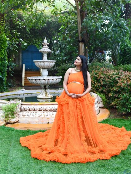 A grand portrait of a mom-to-be in an orange gown by a garden fountain, looking up peacefully.