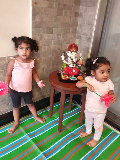 Two little girls standing proudly near the Ganesha idol. They are holding paper flowers they created as offerings during our festive art class.