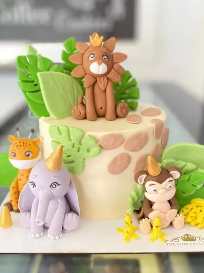 A cute jungle party cake with a crowned lion, a playful monkey, a purple elephant, and a cheetah. The fondant animals are all wearing party hats.