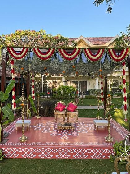 A traditional Brahmin wedding mandap set in a lush garden. The structure is decorated with red and white floral garlands, banana plants, and a hand-painted kolam on the floor.