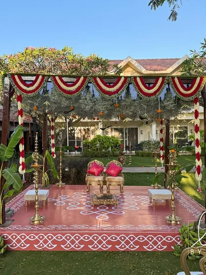 A traditional Brahmin wedding mandap set in a lush garden. The structure is decorated with red and white floral garlands, banana plants, and a hand-painted kolam on the floor.