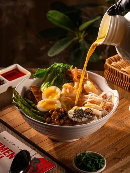 A rich broth being poured into a bowl of ramen. The steam, action, and abundance of toppings make this an incredibly appetizing shot.