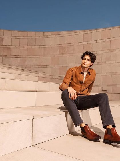 A relaxed, seated pose from the same shoe campaign. This image showcases a different style of boot, with the styling and setting conveying a sense of modern, effortless cool.