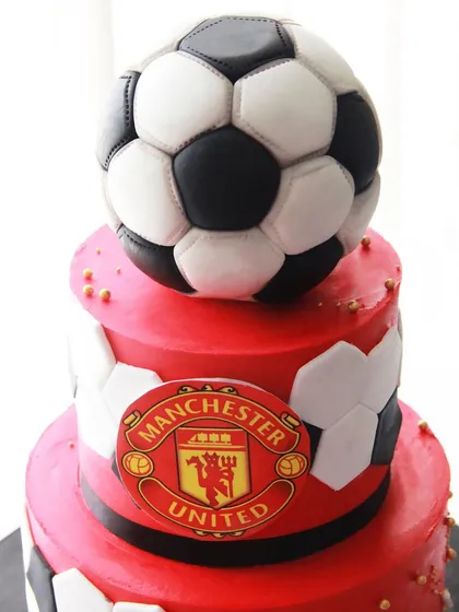 A close-up of the Manchester United football cake, showing the iconic red color and club crest. The football on top is completely edible.