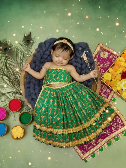 A full setup for a baby Radha shoot, with a green lehenga, peacock feathers, and a box of colorful flowers.