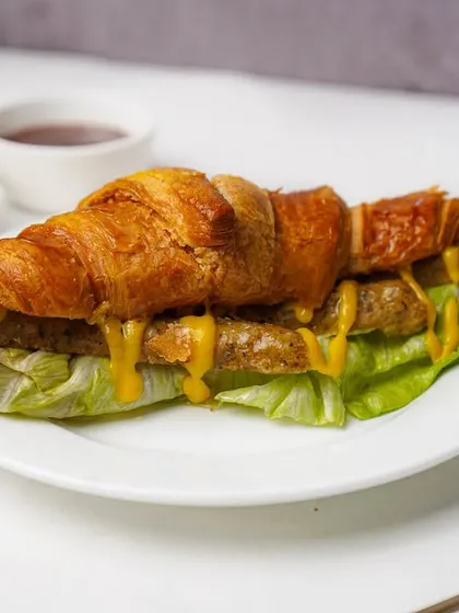 A sausage and lettuce croissant sandwich with a generous drizzle of yellow mustard, presented on a white plate. The simple props keep the focus on this delicious cafe-style offering.