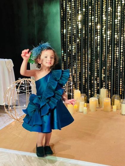 The "Desire" dress in a deep teal blue. The rich color combined with the dramatic ruffles and a matching feathered headpiece creates a truly unique and glamorous party look.