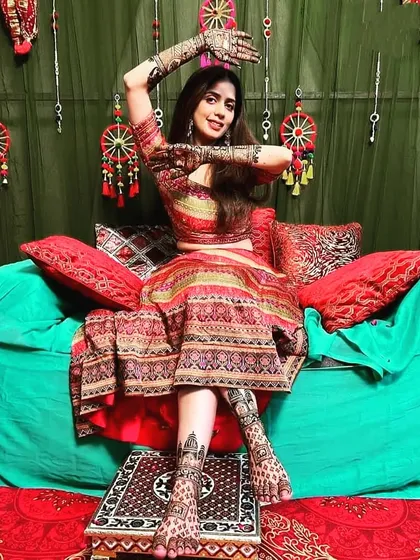 A happy bride celebrating her mehndi function. She is showcasing her full bridal set, with detailed, matching designs on both her hands and feet.