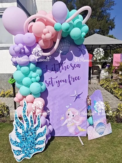 A mermaid-themed photo prop for an outdoor party. The sign reads "Let the sea set you free" and is decorated with pastel balloons and a glittery coral cutout, perfect for a sunny celebration.