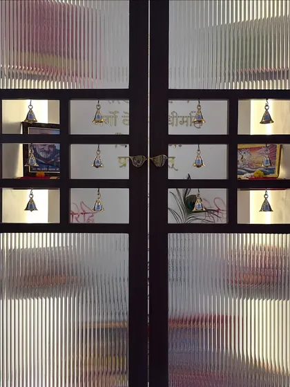 A stunning pooja room door made of dark wood and fluted glass, with small square openings adorned with traditional brass bells.