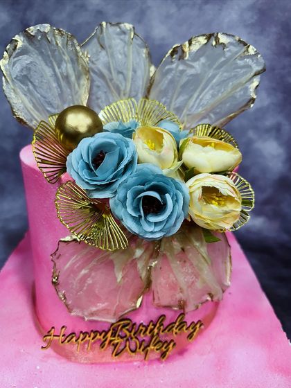 A vibrant pink cake adorned with delicate rice paper sails and beautiful blue and yellow sugar flowers. The gold accents add a touch of glamour to this artistic birthday design.