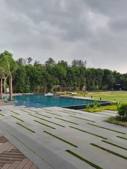 The expansive swimming pool of a Delhi farmhouse, featuring a modern design with a wooden deck and stone pavers with grass joints. The view extends across a vast lawn, creating a sense of openness and luxury.