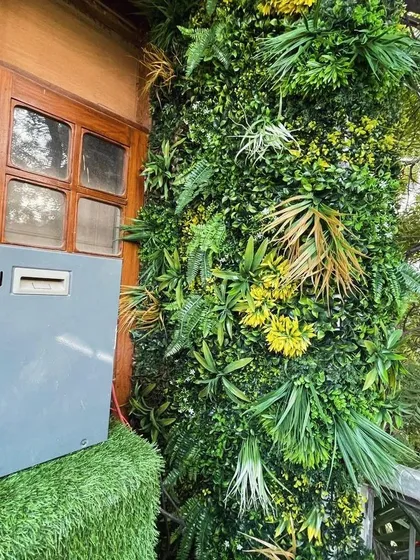 An artificial vertical garden installed on an exterior wall next to a wooden door. This installation effectively hides utility boxes and adds a lush, green texture to the building's entrance.