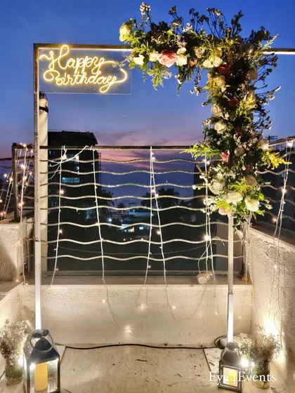 For this terrace birthday party, I created a modern and chic atmosphere with a custom neon "Happy Birthday" sign on a floral arch, set against a beautiful twilight city view. The setup includes fairy lights and lanterns for a warm, inviting glow.