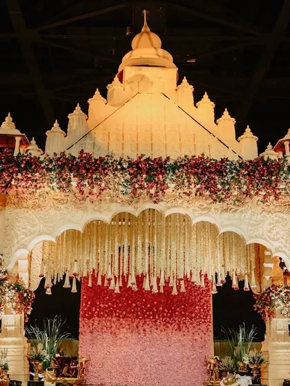 This mandap features a backdrop of cascading red and pink roses, creating a stunning floral wall for the ceremony. The structure itself is inspired by traditional Indian architecture.