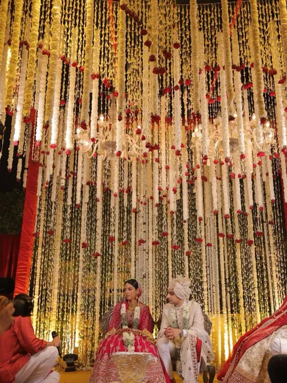 A floral haven for a sacred union. This mandap features an ethereal cascade of red roses and delicate white floral strings, with chandeliers casting a warm glow as the couple partakes in the wedding rituals.