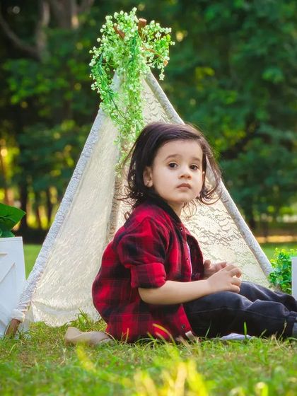 A toddler sits by a lace teepee in a park, looking thoughtful. This outdoor setup adds a touch of bohemian charm to the portrait.