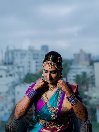 A beautiful portrait of a bride adjusting her necklace, with the Mumbai skyline visible through the window. A blend of tradition and modernity.