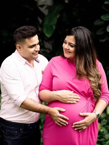 A classic couple's pose against a lush, dark green background. The contrast makes the mom-to-be's bright pink dress stand out, highlighting her and the baby bump beautifully.