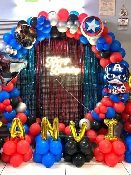 An Avengers-themed birthday party featuring Captain America and Spiderman foil balloons. The setup includes a balloon arch over a fringe curtain and foil letters spelling out the birthday boy's name.