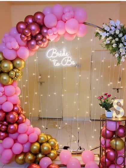 A beautiful half-arch balloon decoration for a bridal shower. The design uses pink, magenta, and gold chrome balloons, with a 'Bride to be' neon sign and a curtain of fairy lights for a magical glow.