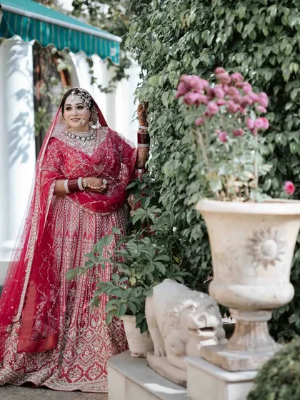 A classic bridal portrait. The bride, in her beautiful red lehenga, poses gracefully in a garden setting, creating a timeless and elegant image.