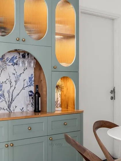A close-up of a beautifully crafted crockery unit, featuring sage green cabinetry with curved details and fluted glass. The arched niche with bird-patterned wallpaper adds a whimsical and artistic touch to this functional piece.