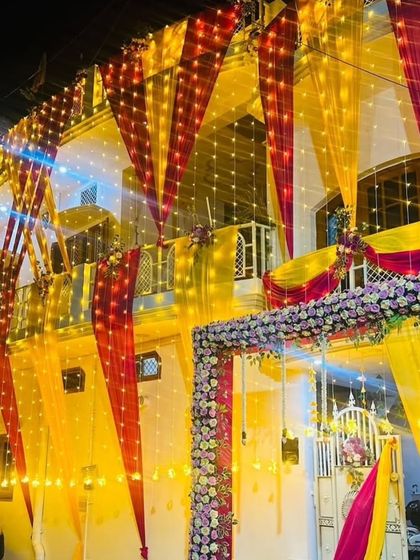 A beautiful night view of a wedding house entrance. The gate is decorated with a floral arch, and the house is lit up with yellow and red drapes and lights.