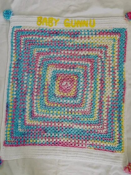 A full view of the "Baby Gunnu" blanket laid flat. The granny square pattern creates a beautiful spiral of pastel colors, and the custom name makes it a cherished keepsake.