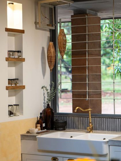 The kitchen sink area, featuring a ceramic farmhouse sink and brass fixtures. The window provides a direct view of the mango trees, making daily chores a more pleasant experience. Open shelves are made from upcycled wood.