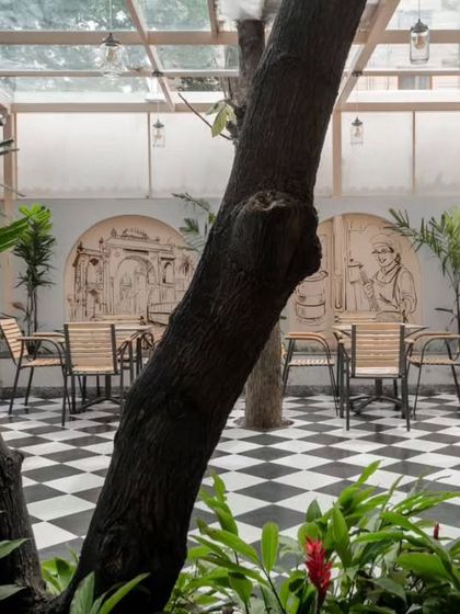 The outdoor seating area is nestled among existing teak and mango trees. A large mural adds an artistic touch to the boundary wall.