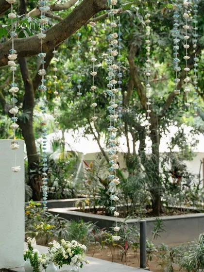 Delicate floral strings in shades of blue and white hang from the trees at the Rishikesh Mehndi, adding a touch of whimsy and movement to the serene outdoor setting.