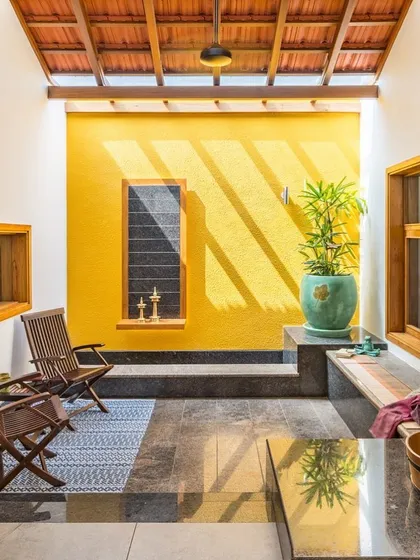 A sun-drenched courtyard featuring a bold yellow wall. The overhead skylight casts dramatic shadows that move across the space, highlighting the texture of the wall and the simplicity of the seating.