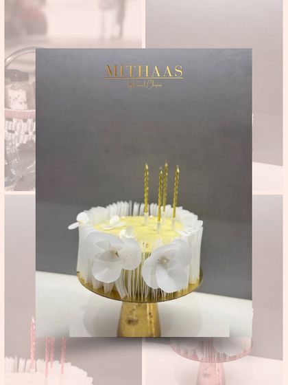 An elegant and minimalist celebration cake. This one is adorned with delicate white edible flowers and shimmering gold candles, presented against a sophisticated grey backdrop.