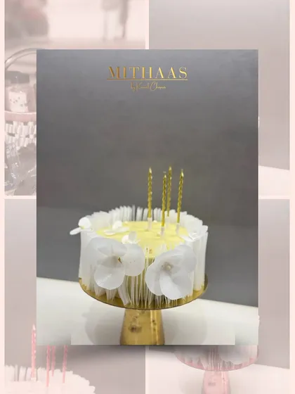 An elegant white cake adorned with delicate, wafer-thin white floral details around its base. The shimmering gold candles add a touch of warmth and celebration to this minimalist design.