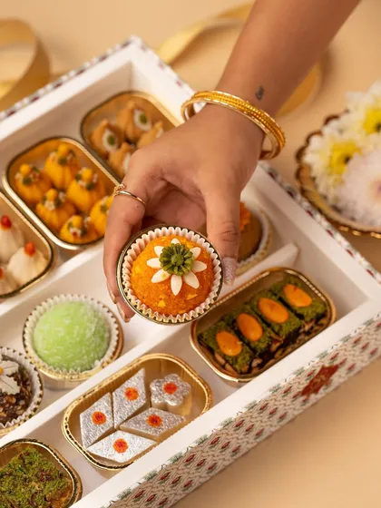 A box of assorted Indian sweets is a feast for the eyes. This shot highlights the vibrant colors and intricate details of each piece, with a hand selecting one to create a sense of interaction and desire.