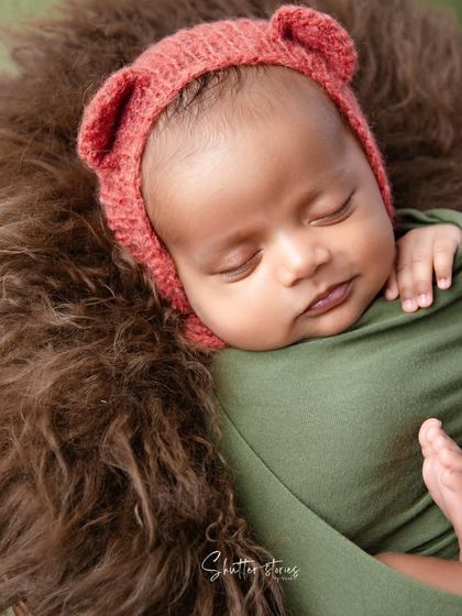 A cozy, earth-toned portrait with a baby in an olive green wrap and a cute bear bonnet, nestled in a brown fur-lined basket.