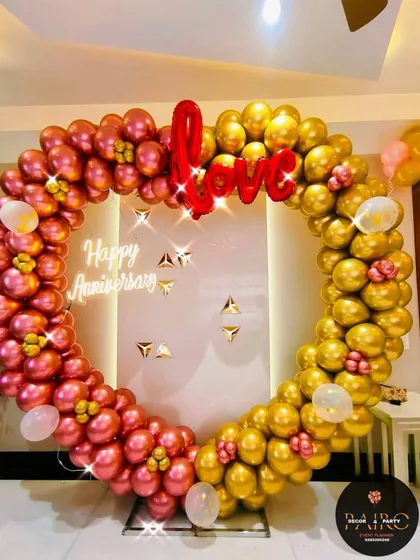 A stylish heart-shaped balloon backdrop for an anniversary. This design uses metallic rose gold and gold balloons, a "Love" foil balloon, and a central "Happy Anniversary" neon sign for a modern look.