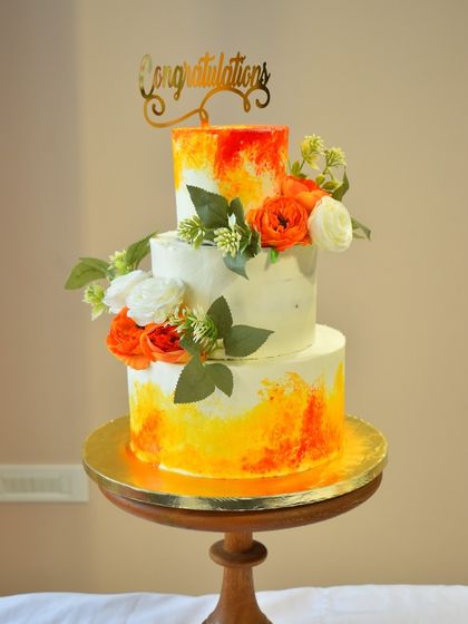 This three-tier cake uses a vibrant watercolor technique with sunset orange and yellow hues. Adorned with fresh flowers and a "Congratulations" topper, it's a bright and cheerful centerpiece for a wedding or engagement party.