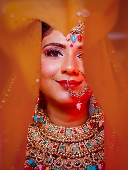 A playful peek through the dupatta. This close-up shot is full of life, highlighting the bride's vibrant smile, colorful jewelry, and the joyful energy of the wedding day.
