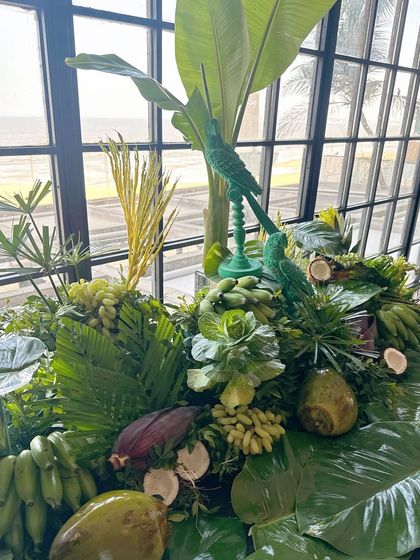 A tablescape covered in banana leaves, coconuts, and other tropical fruits and foliage, with green parrot figurines adding a playful touch.