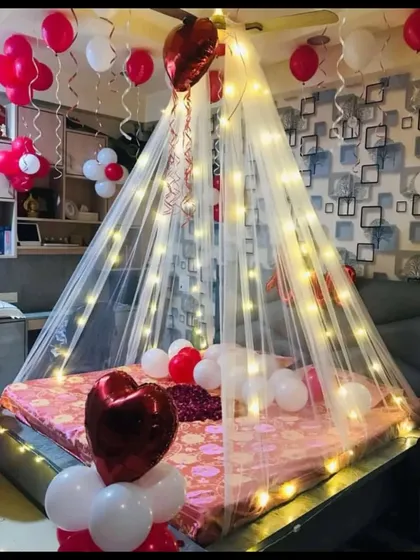 A cozy and romantic corner setup. This decoration features a fairy light canopy over the bed, with red and white balloons, including heart-shaped foils, creating an intimate and warm atmosphere for a special night.