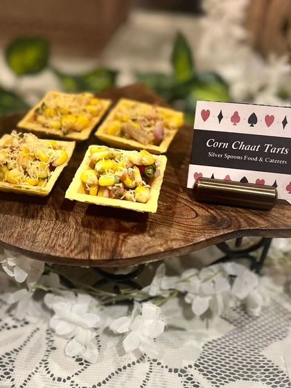 Corn Chaat Tarts presented on a wooden stand with lace and floral details. This bite-sized appetizer combines sweet, savory, and tangy flavors in a crispy tart shell.