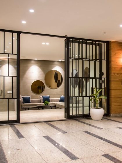 This entryway features industrial-style glass and metal doors that open into a calm, minimalist waiting area. The design uses a simple material palette of wood, stone, and potted plants to create a welcoming first impression.