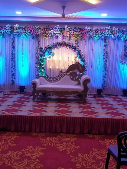 A classic engagement stage setup in a banquet hall. The decor includes a floral arch, a white backdrop with blue uplighting, and a comfortable diwan for the couple, creating a simple yet beautiful setting.