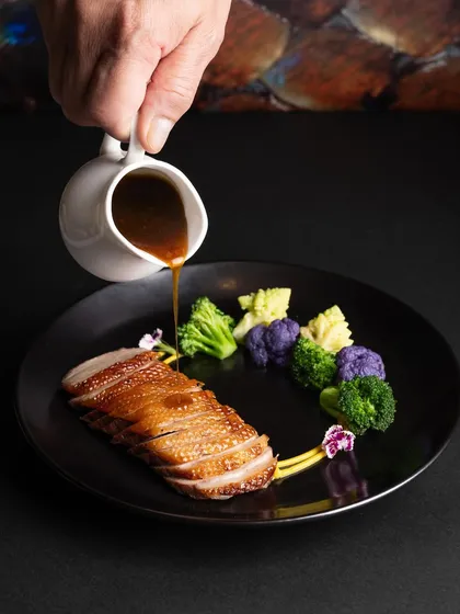 A chef pouring sauce over perfectly cooked duck breast. This action shot captures the culinary process and adds a dynamic, premium feel to the photograph.
